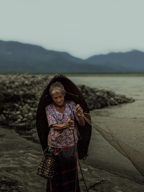 Elderly Woman Fishing Near Scenic River with Mountains in Background