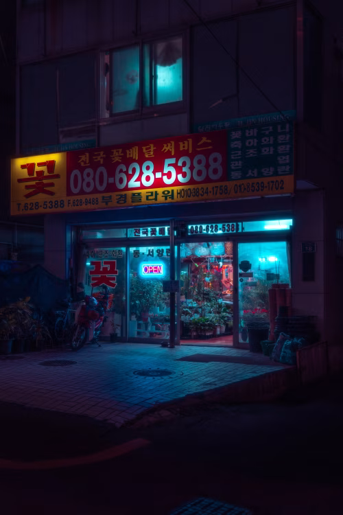 A vibrant city storefront at night, showcasing a colorful and illuminated display with neon signs. The warm glow of the interior and cool street lighting create an atmospheric urban scene.