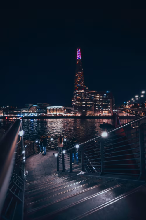 A serene urban night view showcasing a modern skyscraper lit in vibrant colors and a waterfront promenade, creating a contemporary and reflective cityscape scene by the waterside.